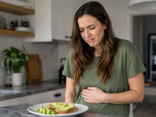 A Western woman holding her stomach in a kitchen while looking uncomfortable beside a plate of avocado toast, suggesting nausea after eating avocado.