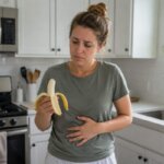 Western woman standing in a bright kitchen, holding a peeled banana while touching her stomach and looking mildly nauseous
