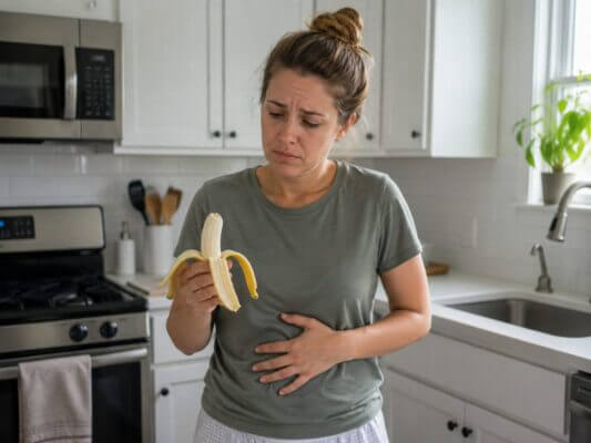 Western woman standing in a bright kitchen, holding a peeled banana while touching her stomach and looking mildly nauseous