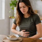 A Western woman sitting at a kitchen table with a bowl of quinoa, gently holding her stomach with a mildly uncomfortable expression after eating.