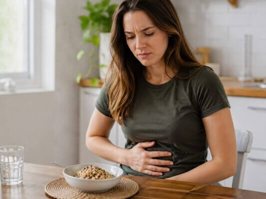 A Western woman sitting at a kitchen table with a bowl of quinoa, gently holding her stomach with a mildly uncomfortable expression after eating.