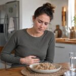 A Western woman sitting at a kitchen table with a bowl of brown rice in front of her, gently holding her stomach with a mildly uncomfortable expression after eating.