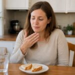A Western woman sitting at a kitchen table with sliced bread and water, looking mildly uncomfortable while holding one hand near her mouth after eating bread.