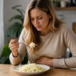A Western woman sitting at a kitchen table with a plate of cabbage salad, gently holding her stomach with an uncomfortable expression after eating.