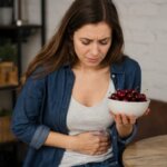 A Western woman holding a bowl of cherries while clutching her stomach in a kitchen, suggesting digestive discomfort after eating cherries.