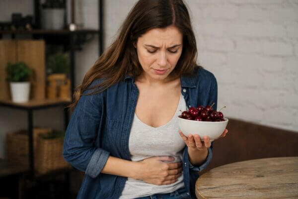 A Western woman holding a bowl of cherries while clutching her stomach in a kitchen, suggesting digestive discomfort after eating cherries.