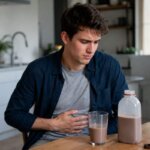 Western man sitting at a kitchen table with chocolate milk, holding his stomach with mild digestive discomfort in a natural photorealistic home setting.