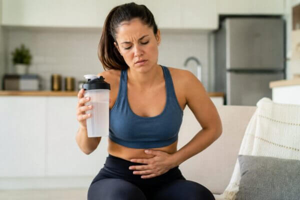 A realistic photo of a Western woman in workout clothes sitting on a couch in a modern kitchen-living space, holding an electrolyte drink bottle while pressing her stomach with a discomforted expression.