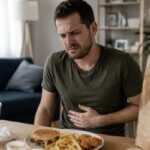 Western man sitting at a table with a burger, fries, fried chicken, soda cup, and takeout bag, holding his stomach with an uncomfortable expression after eating fast food.