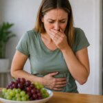 A realistic photo of a Western woman sitting at a wooden table with a bowl of red and green grapes in front of her, holding her stomach with a worried expression near a bathroom doorway.