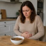 Western woman sitting at a kitchen table holding her stomach with discomfort after eating a bowl of lentils