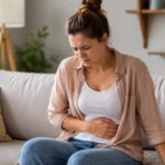 Western woman sitting on a sofa with one hand on her stomach, showing mild digestive discomfort after eating fruit in a natural indoor setting.