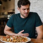 Western man sitting in a casual restaurant with a plate of loaded nachos in front of him, holding his stomach with an uncomfortable expression after eating.