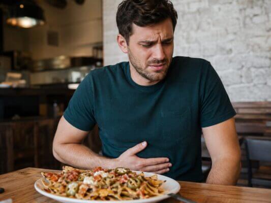 Western man sitting in a casual restaurant with a plate of loaded nachos in front of him, holding his stomach with an uncomfortable expression after eating.