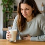 Western woman holding a glass of oat milk at a kitchen table while clutching her stomach with mild digestive discomfort in a natural realistic indoor scene.