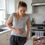 Western woman holding her stomach in a bright kitchen beside a cutting board with sliced raw onions
