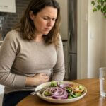 Western woman holding her stomach at a dining table after eating a salad with visible raw red onion slices, showing mild abdominal discomfort in a home kitchen