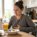 A Western woman sitting at a kitchen table during breakfast, looking mildly nauseous while holding a glass of orange juice, with one hand resting on her upper stomach in a natural photorealistic scene.