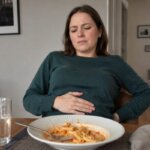 Western woman sitting at a dining table after eating pasta, holding her lower abdomen and looking uncomfortably bloated