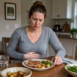 A Western woman sitting at a dining table with a pork meal in front of her, holding her stomach with a mildly nauseous expression in a natural home kitchen setting.