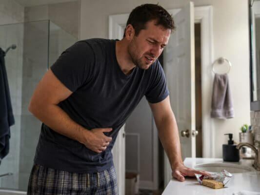 A Western man clutching his stomach in a bathroom after eating a protein bar, showing digestive discomfort in a natural realistic home setting.