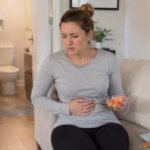 A Western woman sitting on a sofa with a worried expression, holding a small bowl of colorful sugar-free candy while touching her stomach, with an open bathroom door in the background.