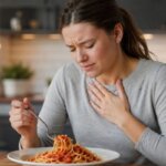 A Western woman holding her chest with discomfort while sitting at a kitchen table with spaghetti in tomato sauce, showing a natural heartburn moment in a realistic home kitchen.