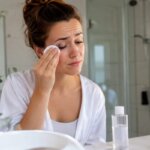 A woman gently removes eye makeup with a cotton pad while experiencing mild burning and irritation.