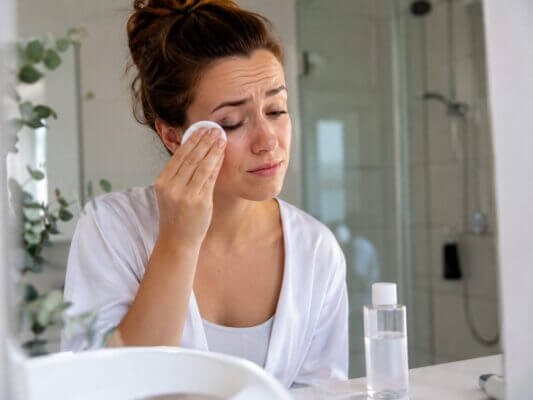 A woman gently removes eye makeup with a cotton pad while experiencing mild burning and irritation.