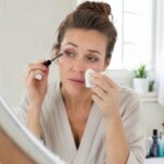 A Western woman applying mascara while gently dabbing a watery eye with a tissue at a bright bathroom vanity.