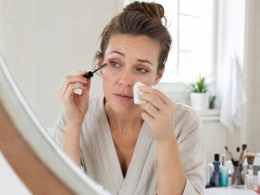 A Western woman applying mascara while gently dabbing a watery eye with a tissue at a bright bathroom vanity.