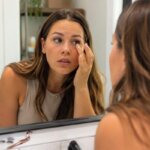 A concerned woman checks her irritated eye in a bathroom mirror after applying false eyelashes.