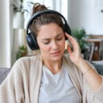 A woman wearing noise-cancelling headphones looks mildly nauseous and uncomfortable while sitting on a sofa at home.