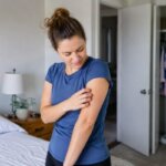 A woman in a blue polyester shirt scratches her itchy upper arm in a softly lit bedroom.