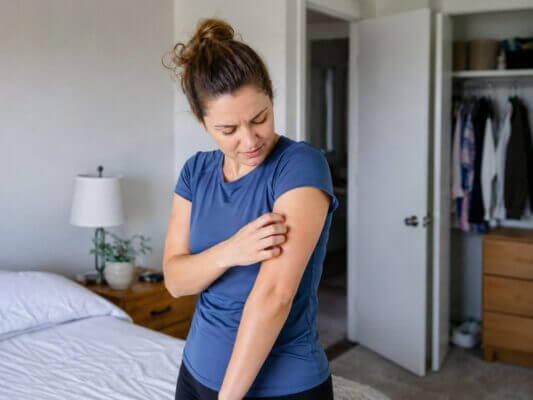 A woman in a blue polyester shirt scratches her itchy upper arm in a softly lit bedroom.