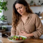 Western woman holding her stomach beside a fresh salad bowl at home, suggesting digestive discomfort after eating salad.