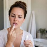 A concerned woman touches her irritated lips while holding a lip balm in a softly lit bathroom.