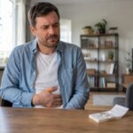 Western man holding his stomach beside an open pack of sugar-free gum at home, suggesting digestive discomfort after chewing xylitol gum.