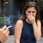 Woman covering her nose and mouth while reacting with nausea and discomfort to nearby cigarette smoke.
