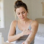 A concerned woman applies lotion to her forearm and notices a stinging sensation in a softly lit bedroom.