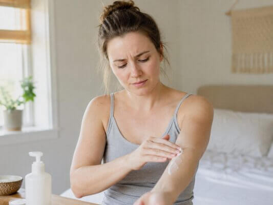 A concerned woman applies lotion to her forearm and notices a stinging sensation in a softly lit bedroom.