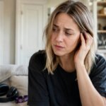 A concerned Western woman touches one ear beside headphones after experiencing muffled hearing from loud music.