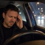 A Western man sitting in a car at night holds his temple in pain while blurred headlights and streetlights glow outside the window.
