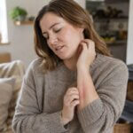 Woman in a wool sweater scratching irritated skin on her neck and forearm indoors